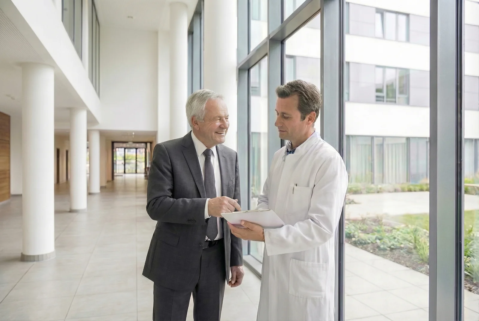 A physician in a white coat reviewing documents with a business consultant in a hospital hallway